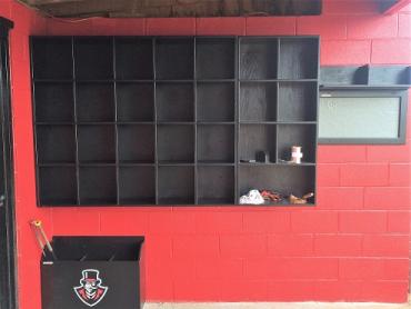 A black wooden 20 Helmet Cubby Rack mounted on a red wall, with individual compartments to store baseball helmets.