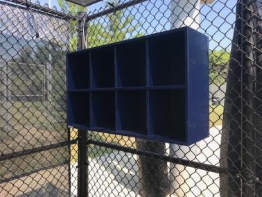 A blue 8 Helmet Cubby Rack mounted on a chain-link fence, designed to hold baseball or softball helmets.