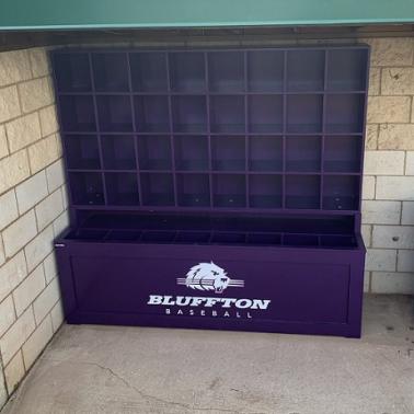A purple dugout helmet and bat storage unit with the Bluffton Baseball team logo on the front and sides. The unit is made of wood and has multiple compartments for helmets and bats.