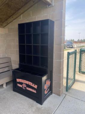A black helmet and bat rack combination unit with multiple compartments, including 16 helmet cubbies, 4 bat compartments, and a coaches storage cubby, displayed outdoors.