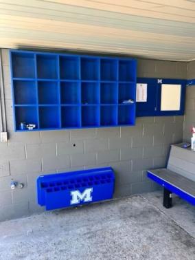 Blue helmet cubby rack installed in a dugout, made of wood with a grey back panel, designed to hold baseball or softball helmets.