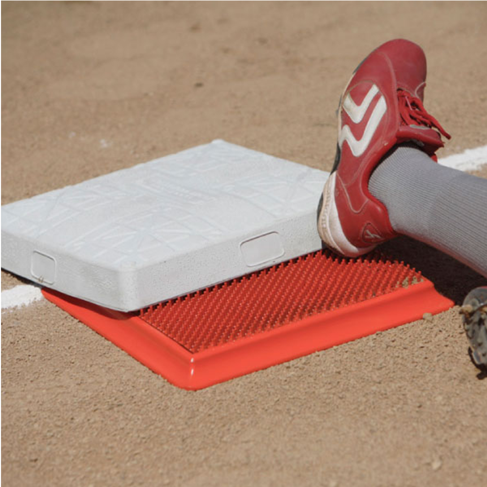 A close-up of a red textured base pad with a white base and a person's leg and red cleat resting on it, set on a dirt field.