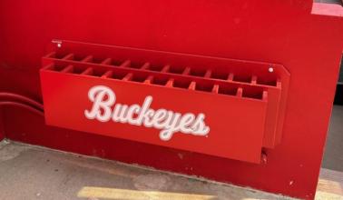 A red vertical bat rack with the word 'Buckeyes' printed on it, mounted on a wall.